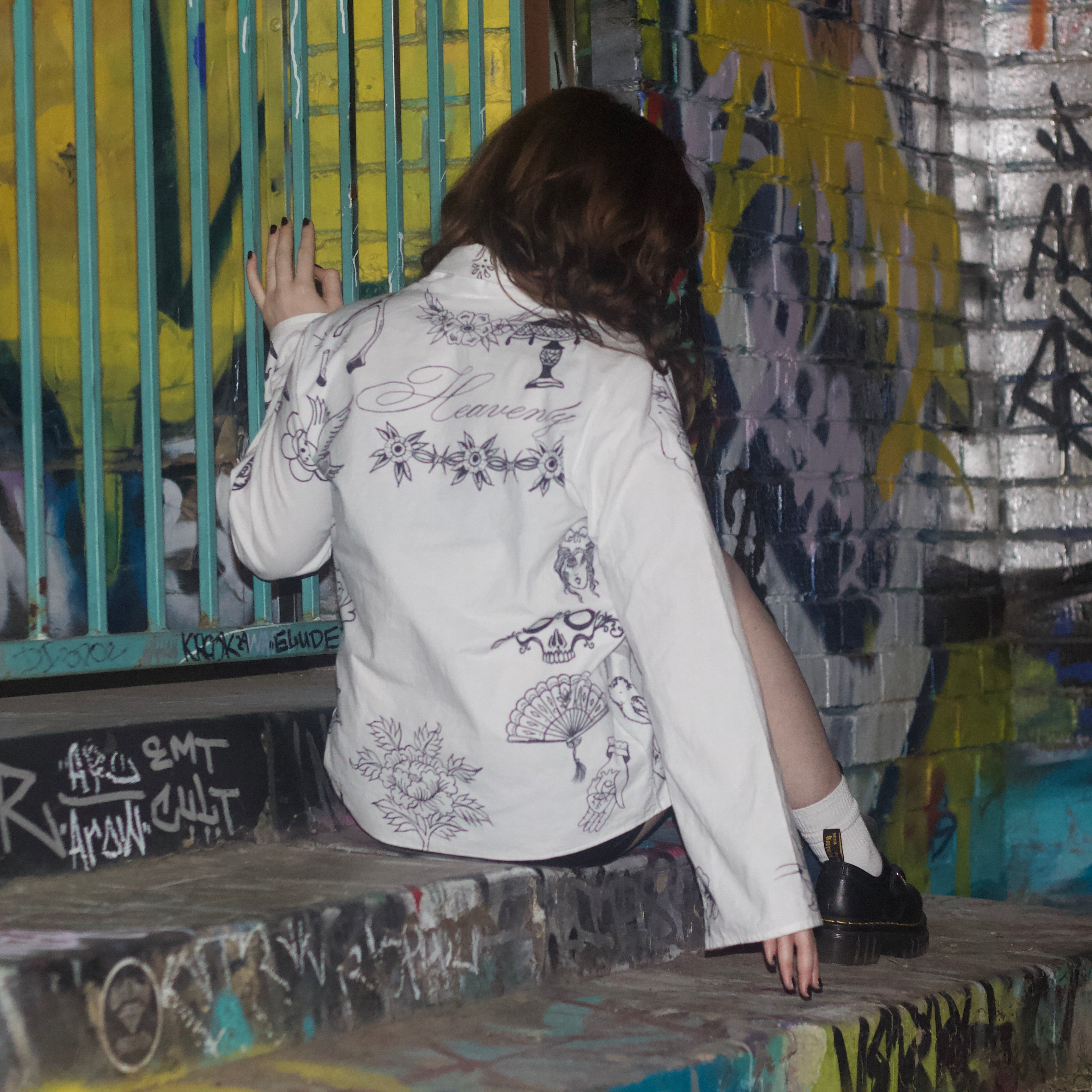 Person wearing a white jacket covered in hand drawn tattoos sitting on a graffiti-covered wall
